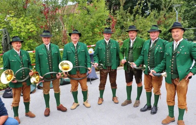 Five men in traditional green and brown attire, holding musical instruments, stand in a row in an outdoor setting.