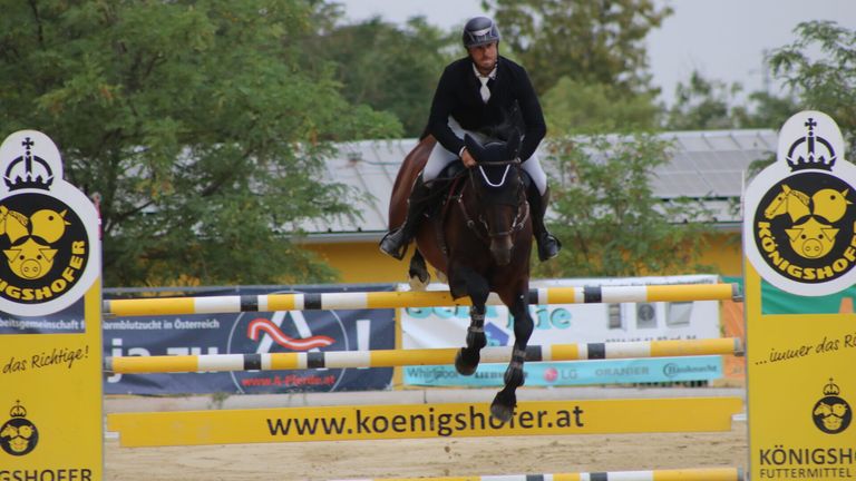 A rider in a black outfit and helmet is on a horse jumping over a hurdle. The horse is brown with black legs. The background shows a blurred banner with text.