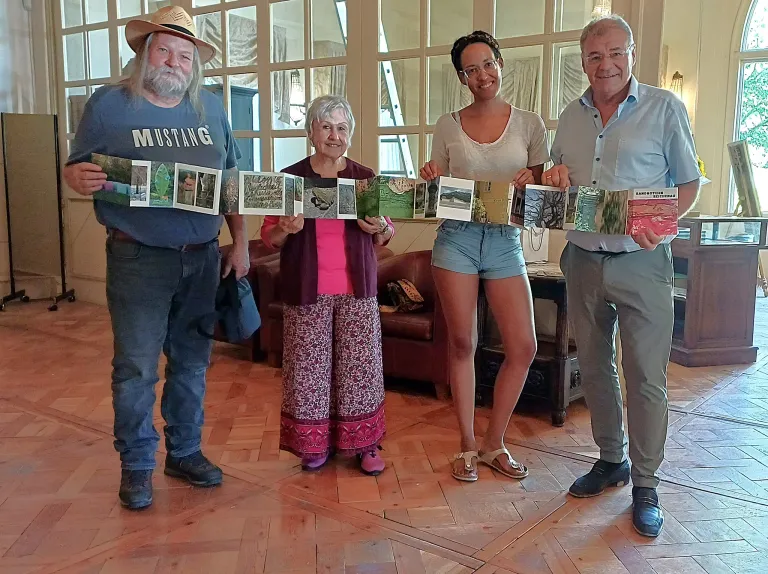 Four people stand in a room, holding a long, folded photo album. The man on the left wears a 'Mustang' shirt. The woman in the middle wears glasses and a pink top. The woman on the right wears denim shorts and sandals. The man on the right wears glasses and a light blue shirt.