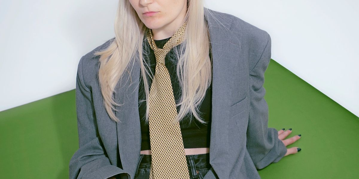 A woman with long blonde hair wearing a black crop top, a gray blazer, and a golden tie with a geometric pattern. She is sitting on a green surface.