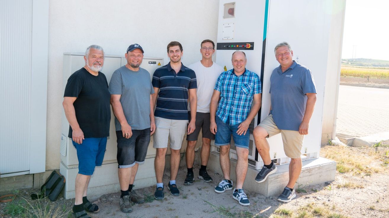 Fünf Männer stehen zusammen vor einem weißen Gebäude mit einem großen Schaltkasten dahinter und lächeln für ein Foto.