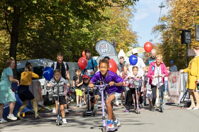 Eine Gruppe von Kindern und Erwachsenen fährt auf Rollern in einem Umzug, einige halten Luftballons, auf einer Straße mit Bäumen und Bannern im Hintergrund.