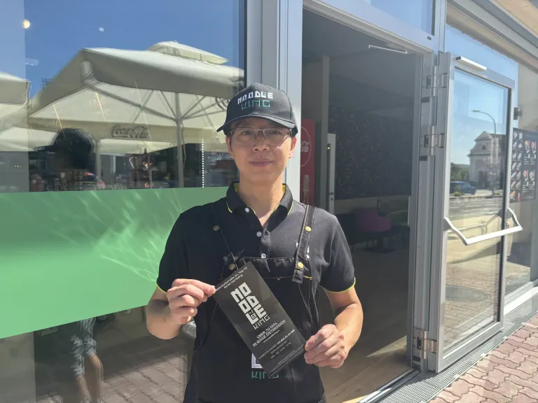 A man in a black apron and cap stands in front of a restaurant, holding a black menu card. The restaurant has a glass door and a Coca-Cola umbrella.