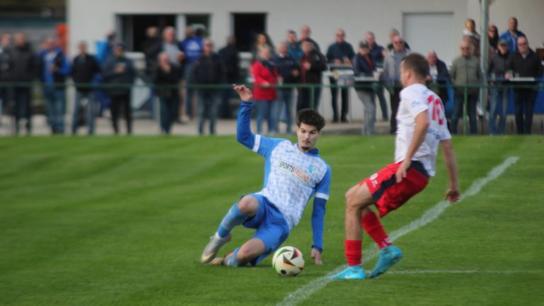 Ein Fußballspieler in blauer Uniform rutscht auf dem Rasenfeld und versucht, den Ball zu schießen, während ein anderer Spieler in roten Shorts verteidigt.