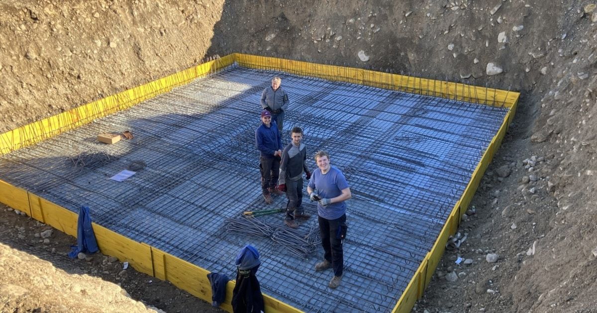 Five construction workers stand on a reinforced concrete slab, wearing gloves and safety gear. The slab is surrounded by yellow barriers.