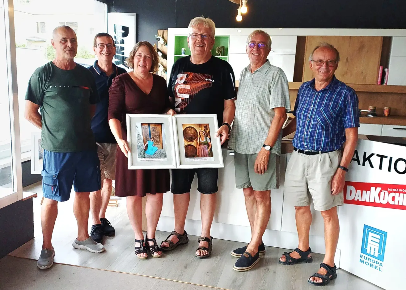Sechs Menschen stehen in einem Raum, lächeln und posieren für ein Foto. Eine Frau hält zwei gerahmte Bilder. Dahinter ist ein weißer Regal mit Gegenständen zu sehen.