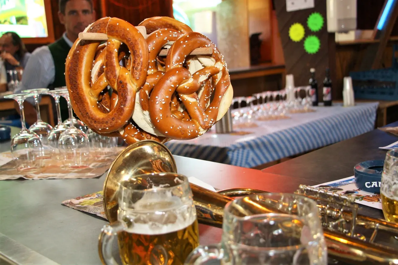 Ein Nahaufnahme von Brezen auf einem Messinghorn mit Gläsern Bier im Vordergrund. Im Hintergrund ist ein Tisch mit Weingläsern und Flaschen gedeckt.