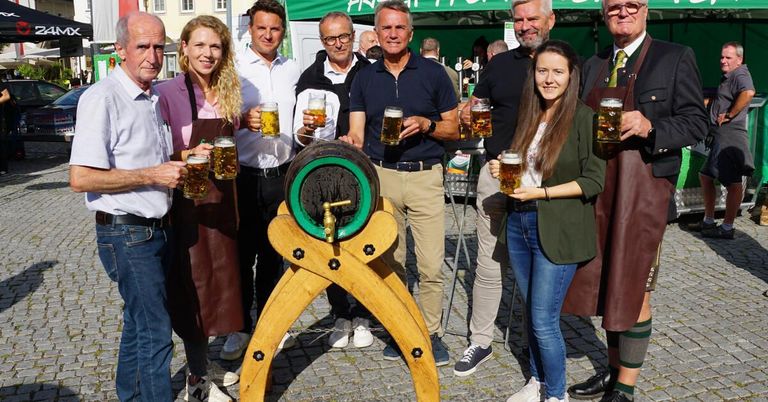 A group of people dressed in traditional attire are posing with beer mugs in front of a large wooden barrel at an outdoor event.