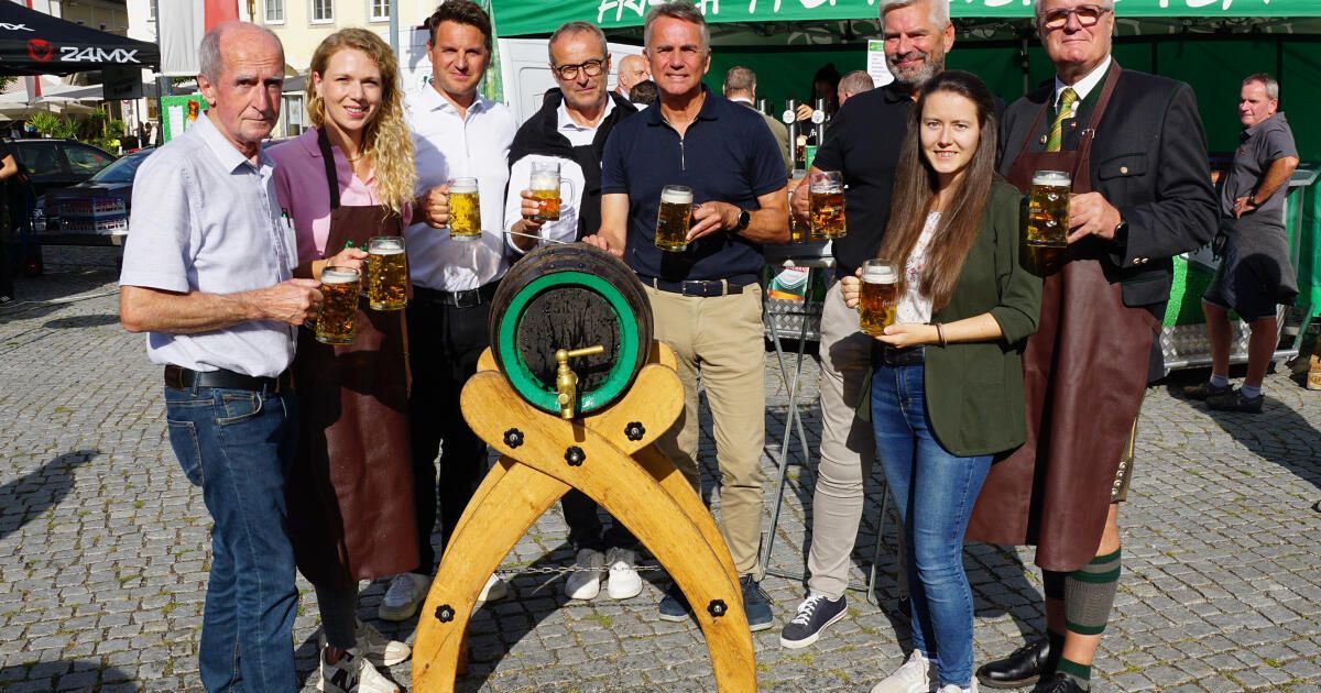 A group of people dressed in traditional attire are posing with beer mugs in front of a large wooden barrel at an outdoor event.