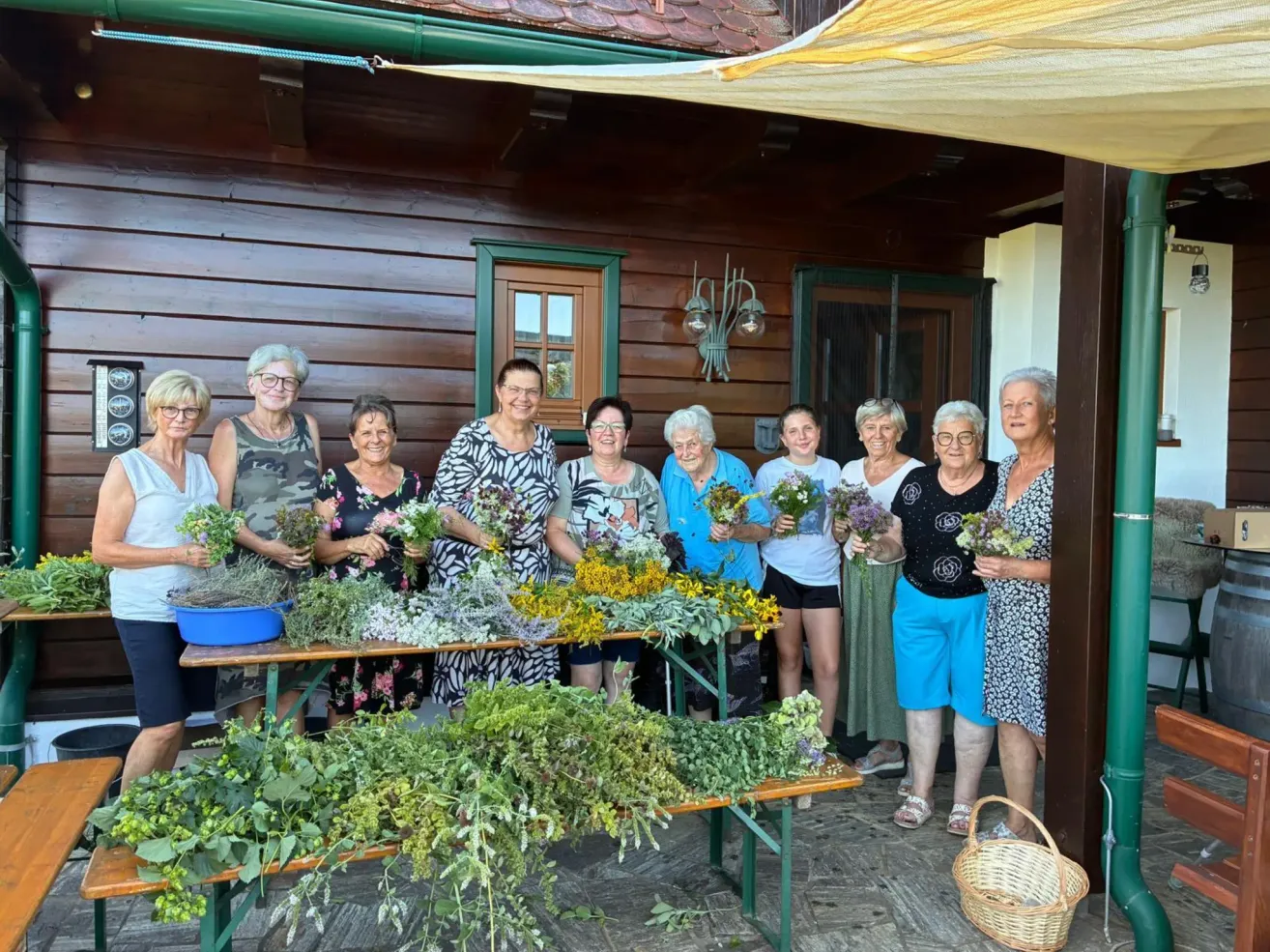Eine Gruppe älterer Frauen steht vor einem Holzhaus und hält verschiedene Blumen und Kräuter in den Händen. Sie lächeln und scheinen für ein Foto zu posieren.