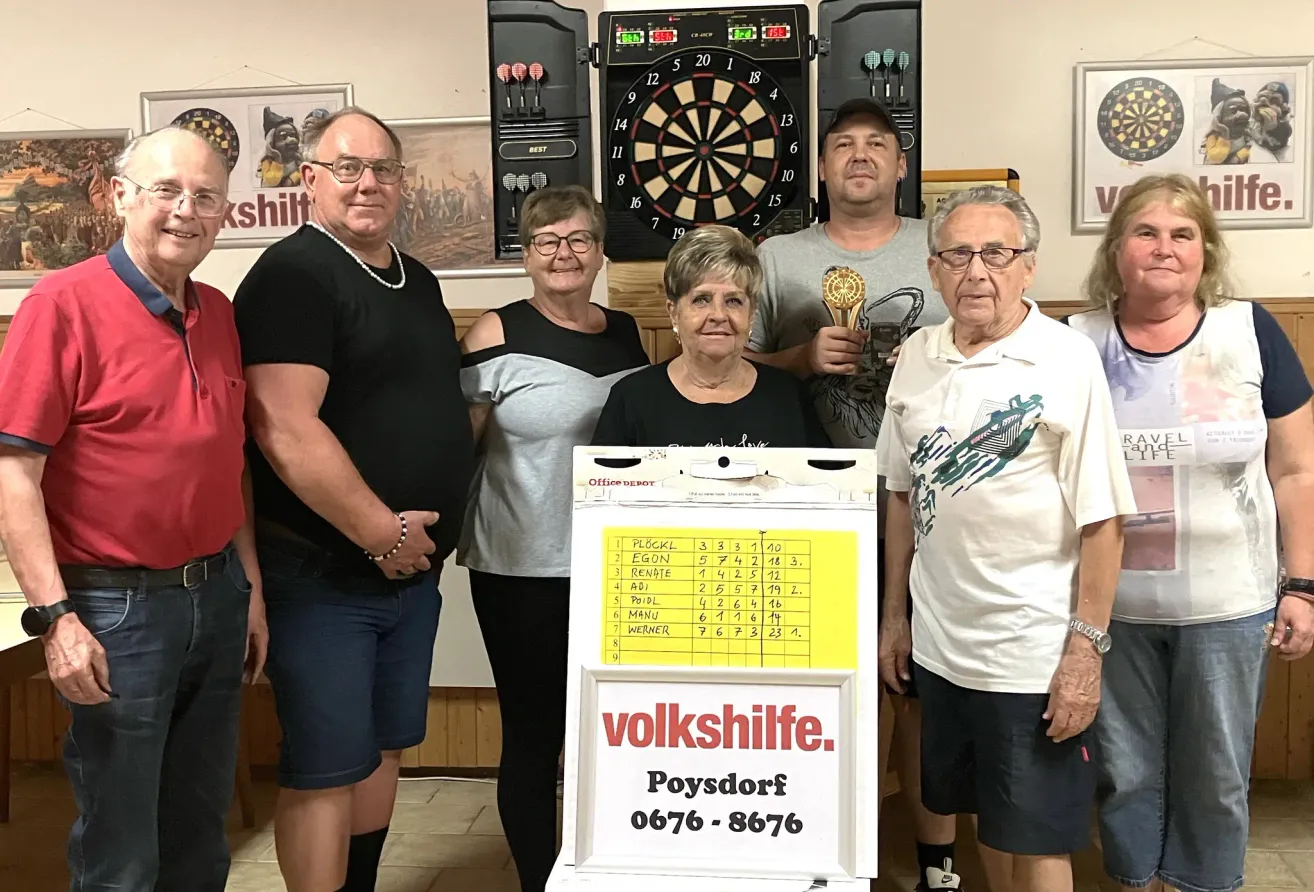 Fünf Personen stehen vor einem Dartboard und einem Punkteanzeiger. Eine Frau in der Mitte hält einen Pokal. Die anderen tragen Freizeitkleidung. Das Dartboard hat die Zahlen 1 bis 20.