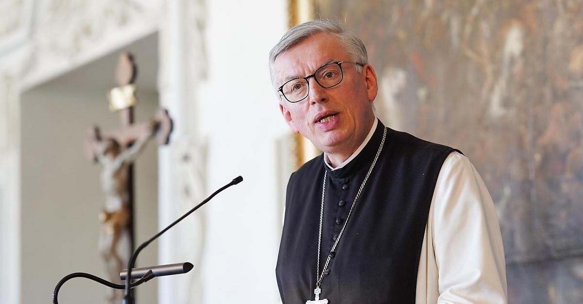 Ein Priester in einem schwarzen Gewand mit Kreuzkette steht an einem Podium mit Mikrofon in einer Kirchenumgebung.
