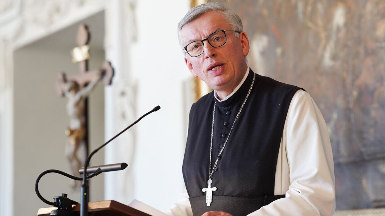 Ein Priester mit Brille und schwarzem Gewand steht an einem Rednerpult und spricht in ein Mikrofon.