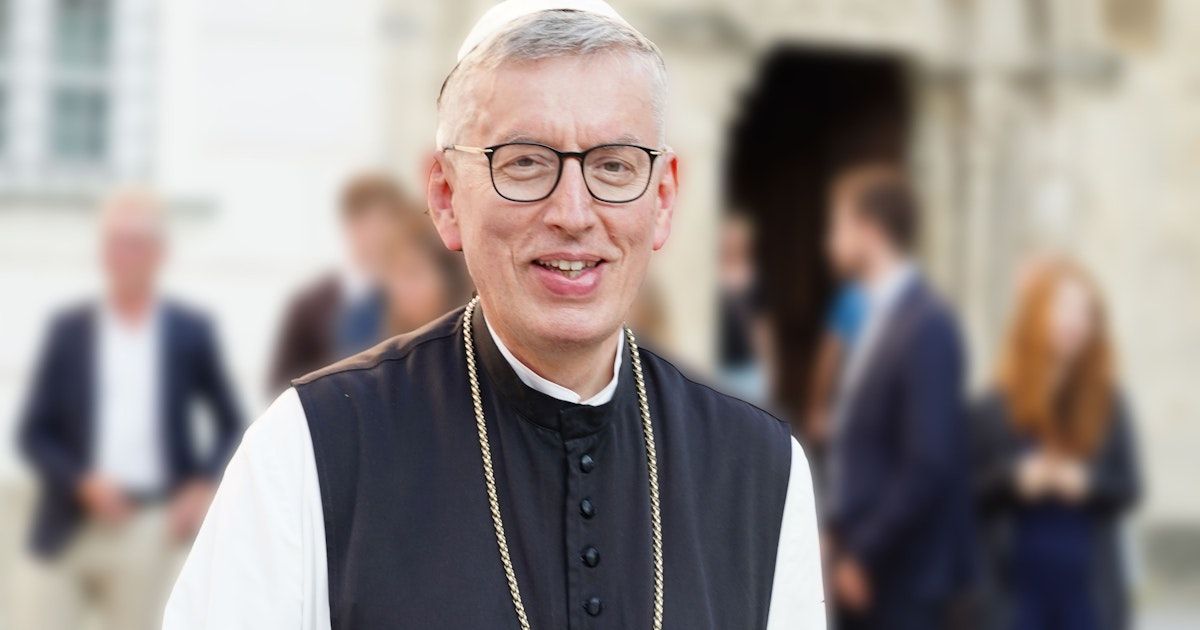 Ein lächelnder Mann mit Brille, einer weißen Mütze und einer schwarzen Weste mit einer goldenen Kette steht vor einem verschwommenen Hintergrund.