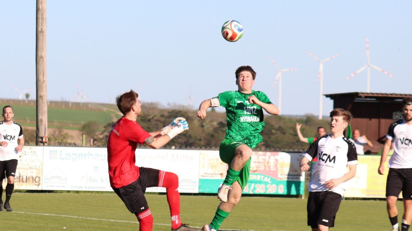 Ein Fußballspieler in Grün springt, um den Ball zu köpfen, während ein anderer Spieler in Rot bereit ist zu blocken. Dahinter eine Mauer mit Werbung und Windrädern in der Ferne.
