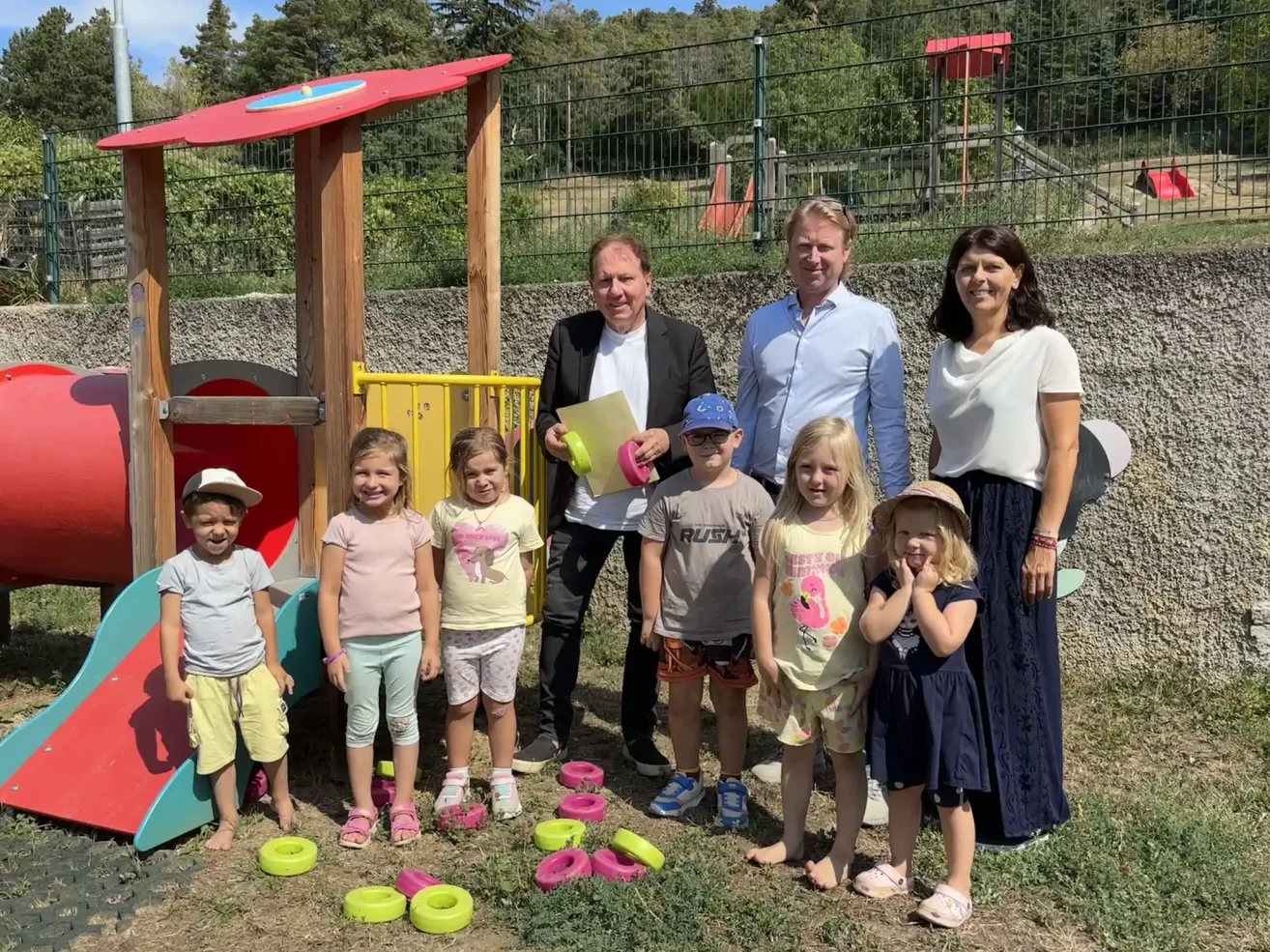 Eine Gruppe von Erwachsenen und Kindern steht vor einem Spielplatz, lächelt und posiert für ein Foto. Die Kinder halten Frisbees und stehen auf einem Rasenbereich. Hinter ihnen befindet sich ein Spielplatz mit einer Rutsche und einem Zaun.