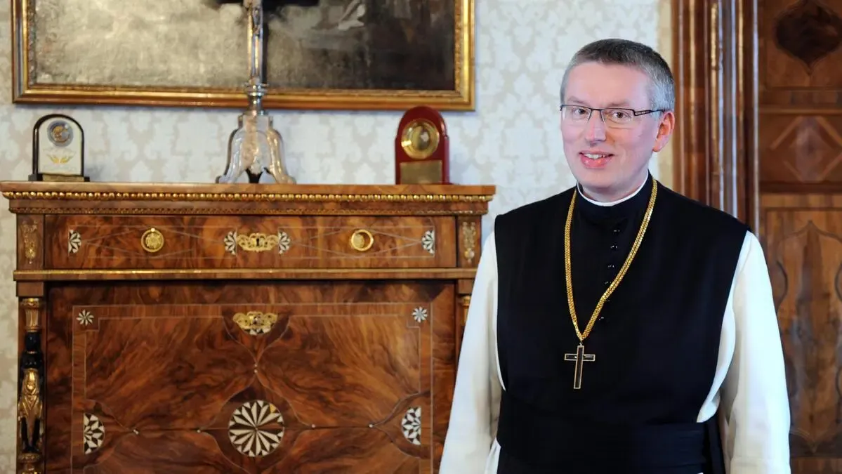Ein Priester steht lächelnd vor einem hölzernen Schrank mit aufwendigen Verzierungen und einem gerahmten Bild darüber. Er trägt eine goldene Kreuzkette.
