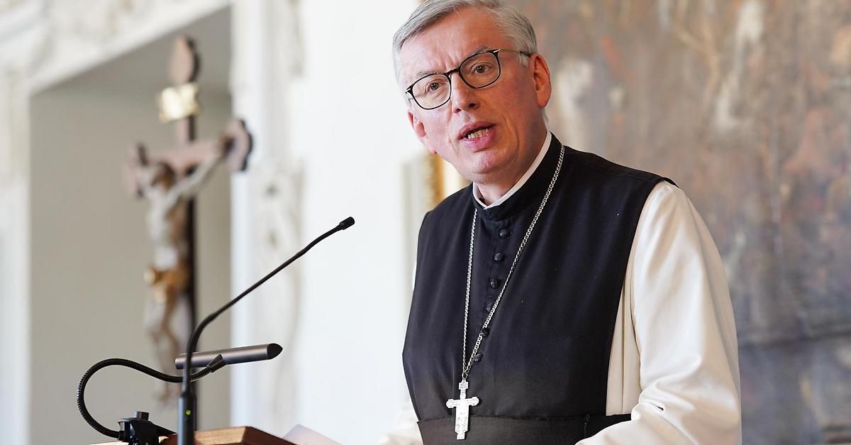 Ein Priester in einem schwarzen Gewand und mit Brille steht an einem Rednerpult mit einem Mikrofon und hält eine Rede in einer Kirche.
