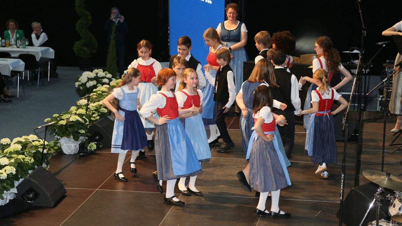 Eine Gruppe von Kindern in traditionellen bayerischen Kostümen führt eine Tanzaufführung auf der Bühne auf, während Erwachsene im Hintergrund zusehen.