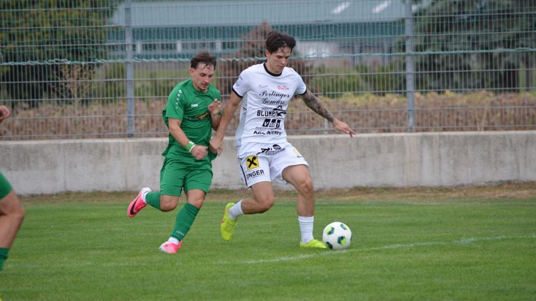 Zwei Fußballspieler in grünen und weißen Trikots spielen auf einem Feld, einer jagt dem Ball hinterher, während der andere verteidigt.