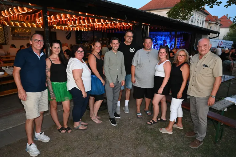 Eine Gruppe von Männern und Frauen steht nachts vor einer Bar und lächelt für ein Foto. Die Bar hat ein Vordach mit Lichterketten.
