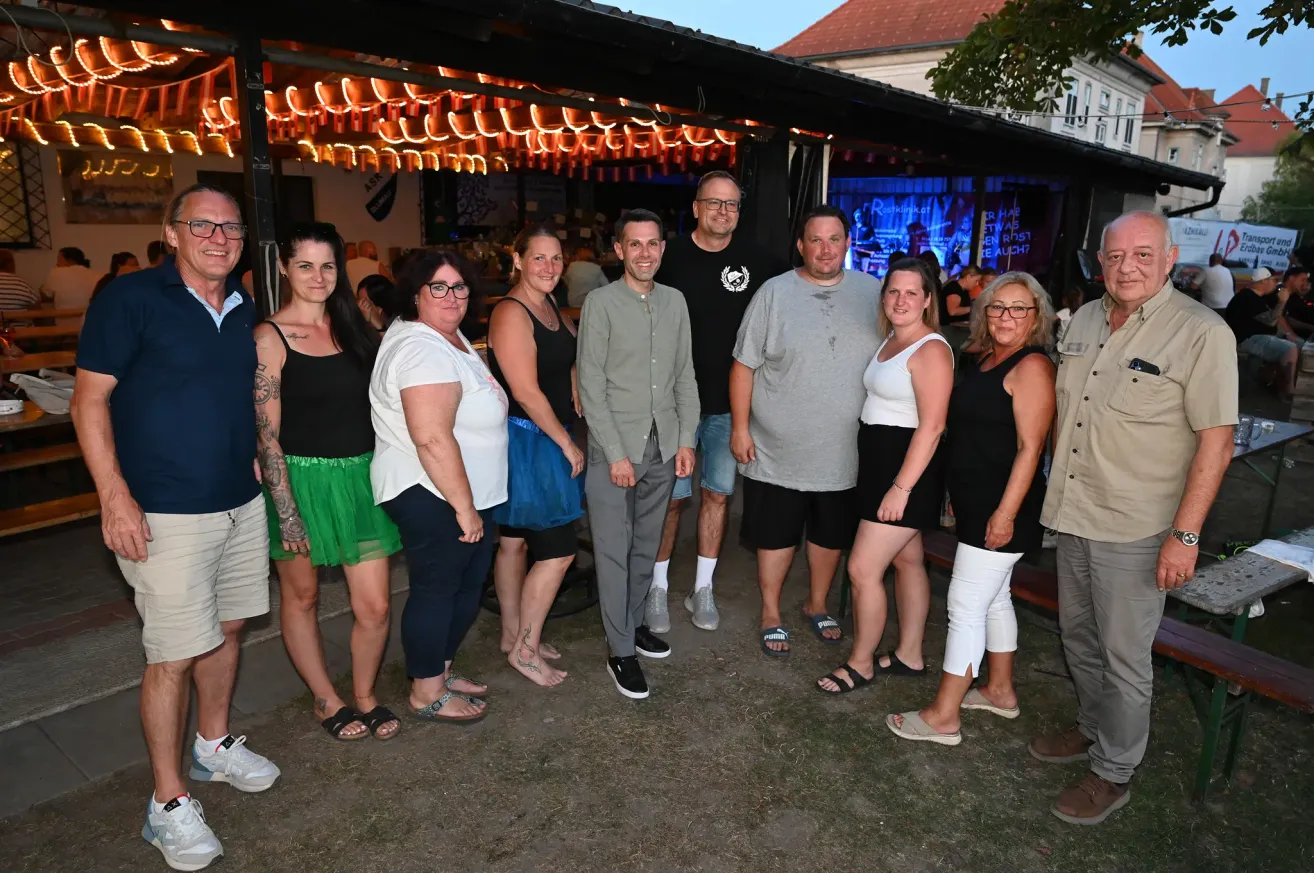 Eine Gruppe von Männern und Frauen steht nachts vor einer Bar und lächelt für ein Foto. Die Bar hat ein Vordach mit Lichterketten.