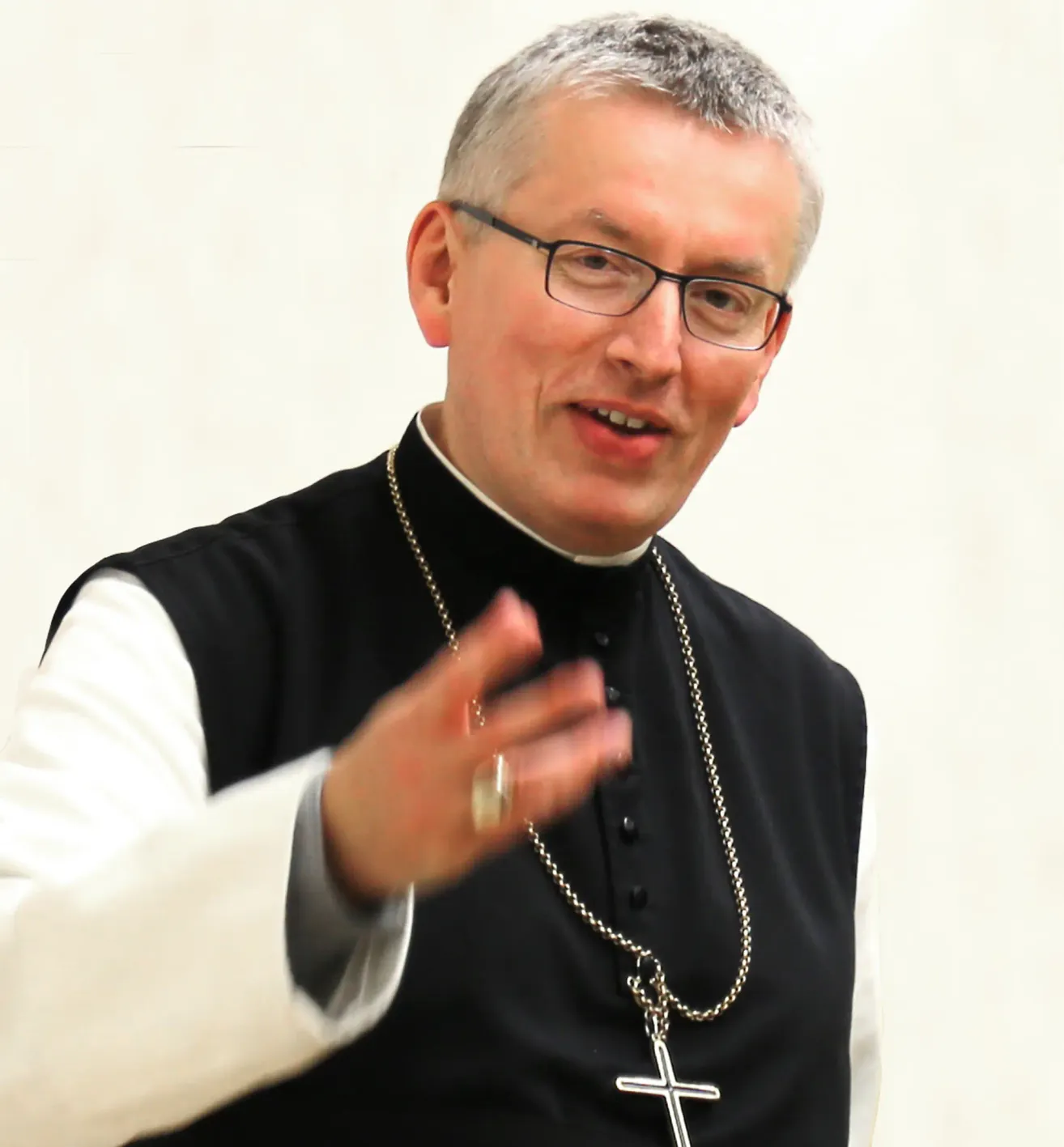 Ein Priester mit grauem Haar und Brille trägt eine schwarze Weste und ein weißes Hemd und gestikuliert mit der Hand.