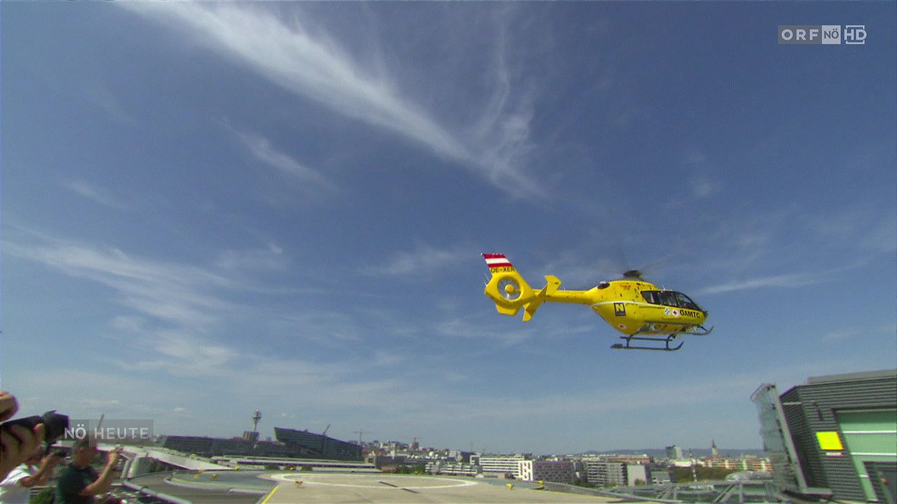 Ein gelber Hubschrauber mit rotem und weißem Heck fliegt über eine Stadt mit Gebäuden und klarem blauem Himmel.