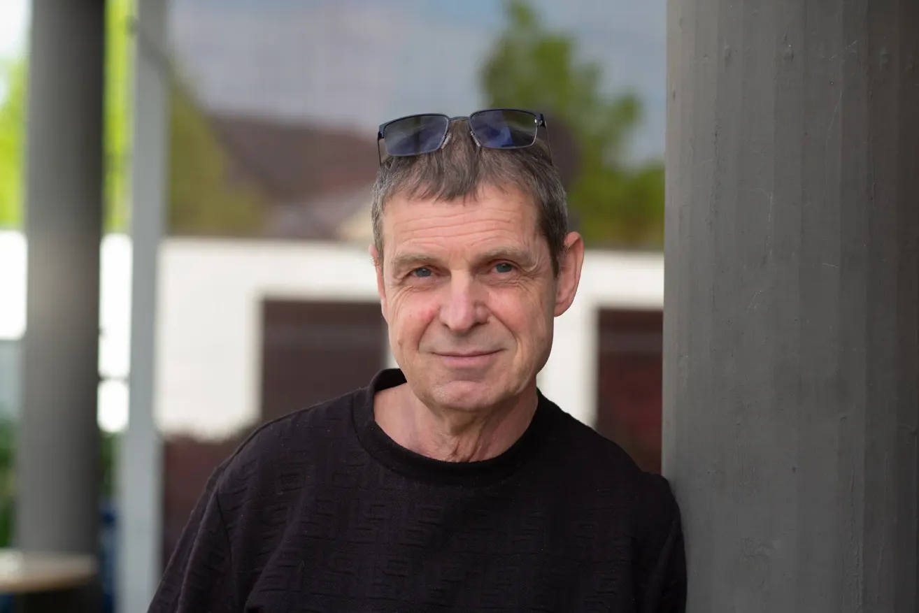 Ein älterer Mann mit grauem Haar, der einen schwarzen Pullover trägt und eine Sonnenbrille auf dem Kopf hat, lächelt und steht draußen.