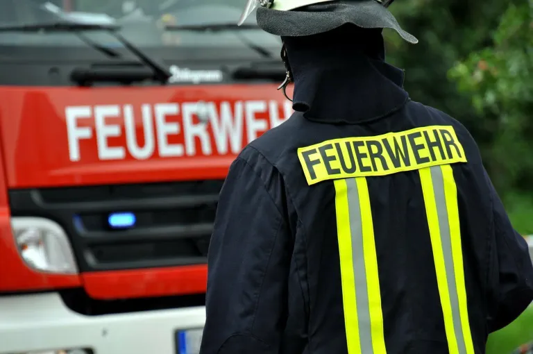Ein Feuerwehrmann steht vor einem roten Feuerwehrauto mit dem Wort 'Feuerwehr' auf der Vorder- und Rückseite seiner Uniform.
