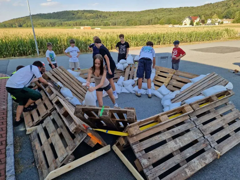 Eine Gruppe von Kindern und Erwachsenen arbeitet zusammen, um eine Struktur aus Holzpaletten und Sandsäcken auf einem offenen Feld zu bauen.