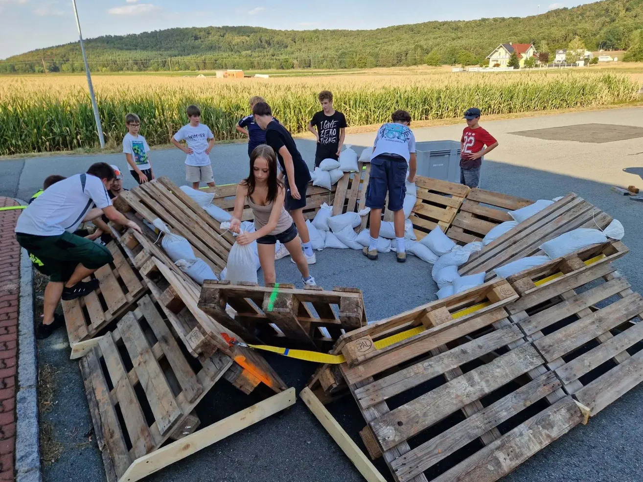 Eine Gruppe von Kindern und Erwachsenen arbeitet zusammen, um eine Struktur aus Holzpaletten und Sandsäcken auf einem offenen Feld zu bauen.