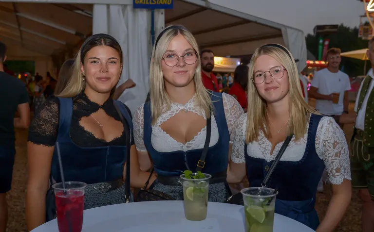 Drei Frauen in traditioneller deutscher Kleidung posieren für ein Foto bei einer Outdoor-Veranstaltung, jede hält ein Getränk mit Limettenscheiben. Im Hintergrund ist verschwommen ein Schild mit der Aufschrift 'GRILLSTAND' und andere Personen zu sehen.