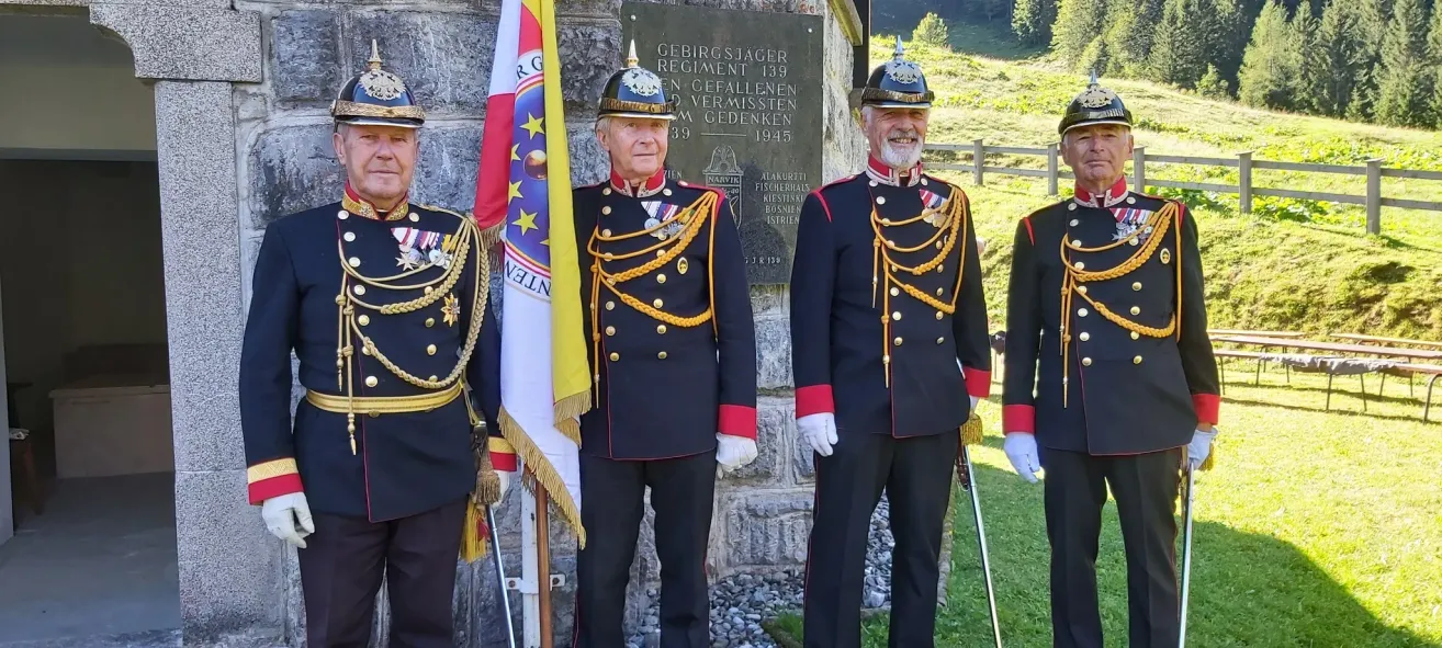 Drei Männer in Militäruniformen stehen vor einem Gedenkstein, einer hält eine Flagge. Der Stein ist mit den Worten 'Gebirgsjäger Regiment 139' graviert.