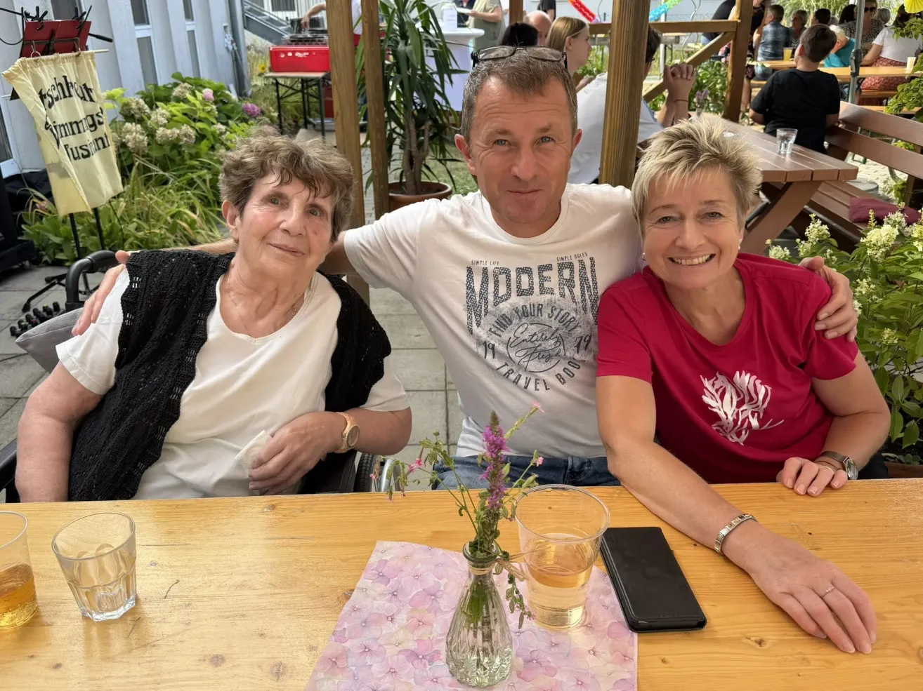 Drei Personen sitzen an einem Tisch in einem Außengarten. Ein Mann mit einem weißen T-Shirt und zwei Frauen, eine mit einem roten Shirt und die andere mit einem weißen Shirt. Eine Vase mit Blumen und ein Glas mit Getränk stehen auf dem Tisch.