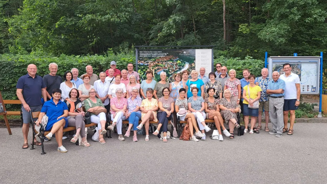 Eine Gruppe von Menschen, wahrscheinlich Touristen, posiert für ein Foto vor einem Schild in einem Park. Sie sitzen auf Bänken und einige stehen.