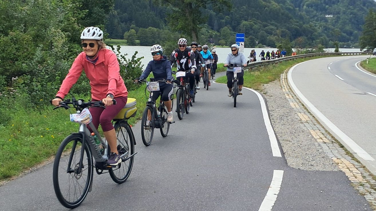 Eine Gruppe von Radfahrern mit Helmen und Radtrikots fährt auf einer Straße am See entlang mit einer malerischen Aussicht auf Berge und Bäume.