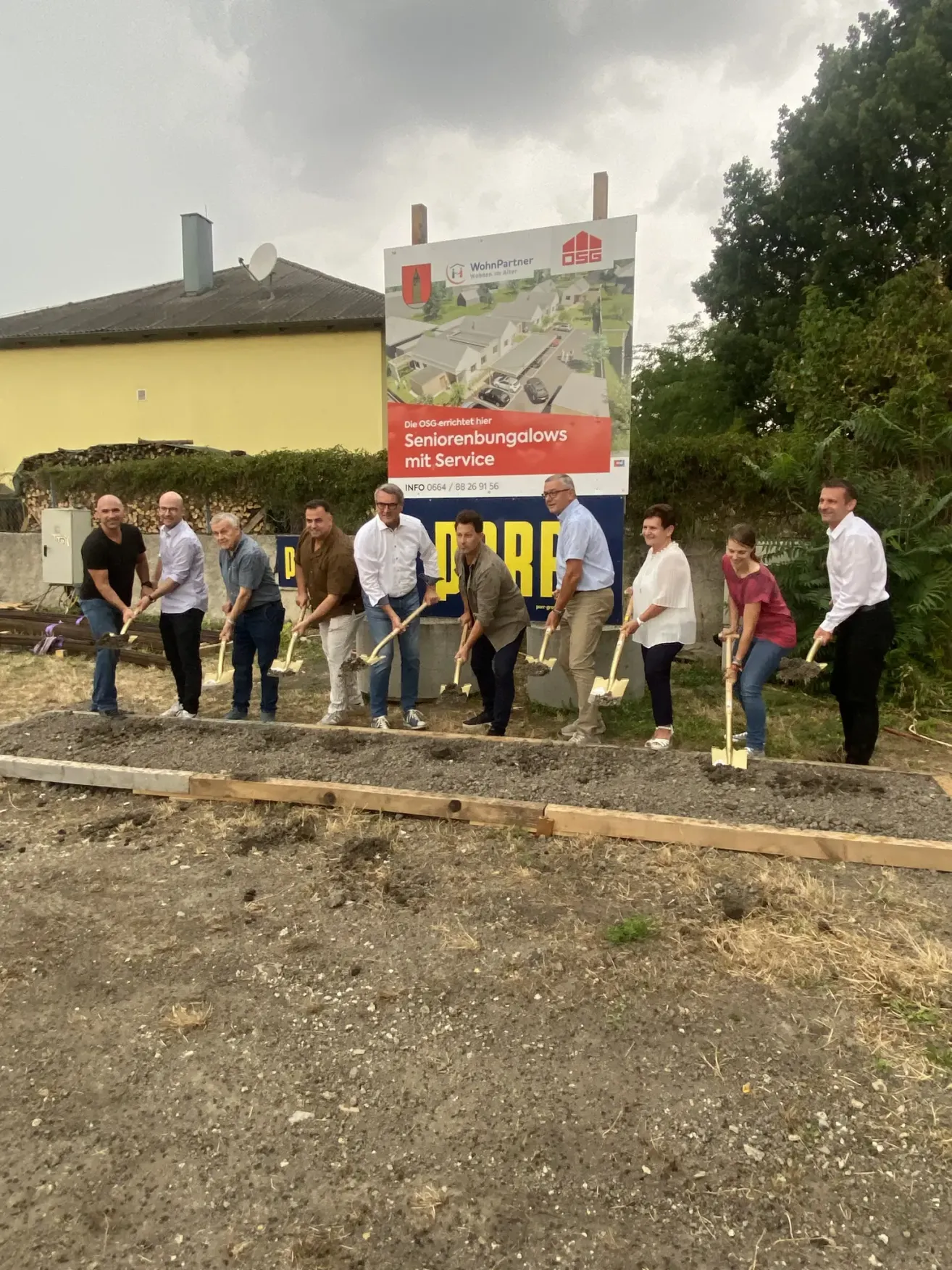 Eine Gruppe von Menschen in formeller Kleidung mit Schaufeln an einer Baustelle, mit einem Banner hinter ihnen, das 'Seniorenbungalows mit Service' besagt.