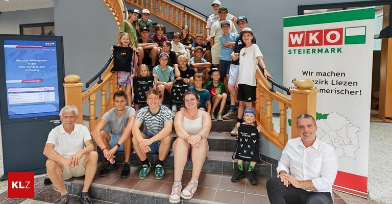 Eine Gruppe von Menschen, einschließlich Kindern und Erwachsenen, posiert für ein Foto auf einer Treppe. Sie tragen alle Mützen und einige halten Taschen. Die Treppe hat Holzgeländer. Ein Schild mit dem Text 'WK STEIER' befindet sich auf der rechten Seite.