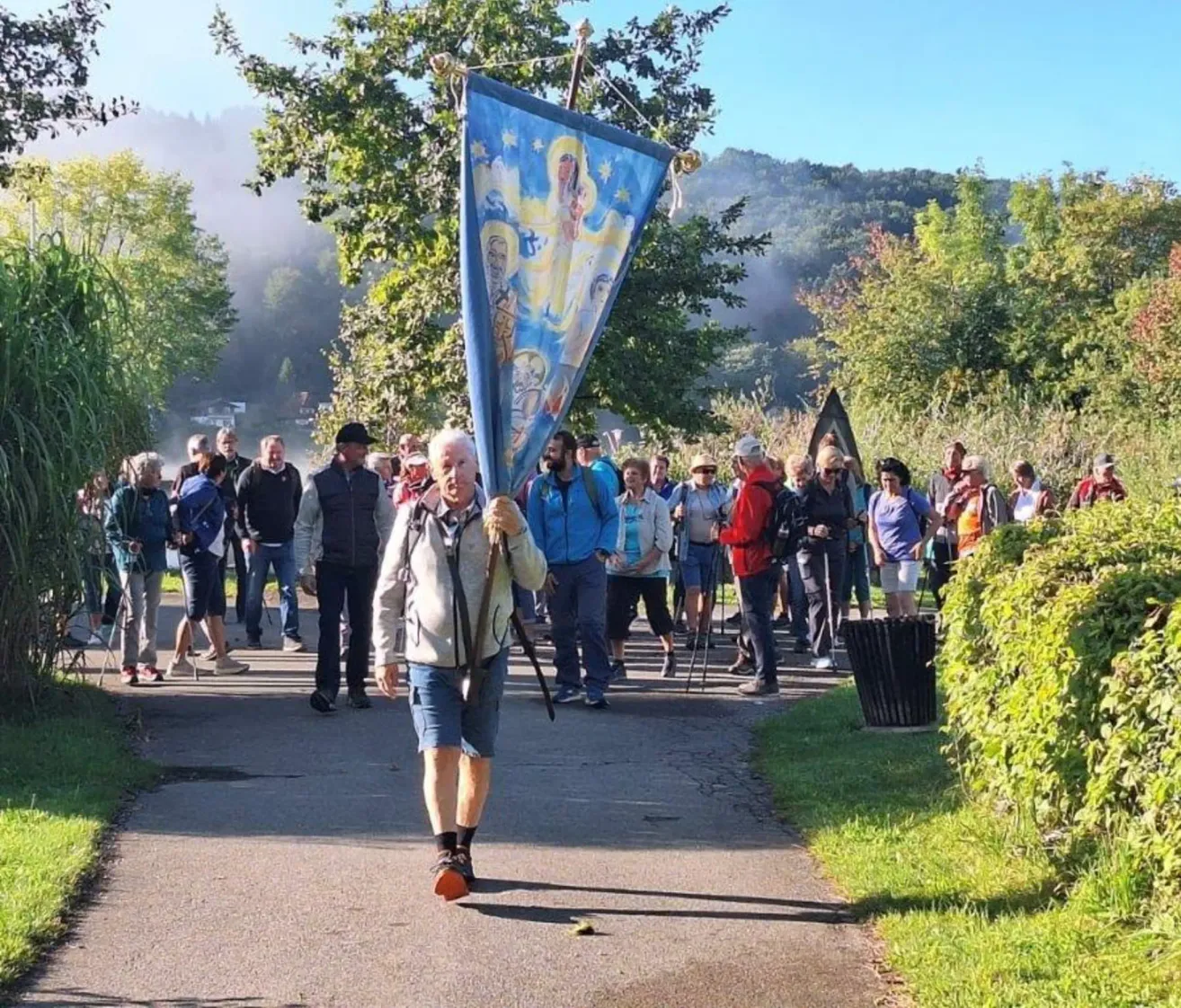 A group of people are walking on a pathway, one man is carrying a blue banner with a religious image.