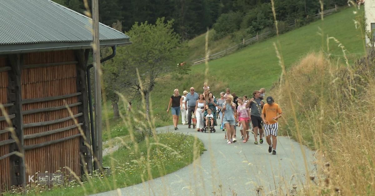 Eine Gruppe von Menschen, darunter Erwachsene und Kinder, geht auf einem Schotterweg in einem ländlichen Gebiet. Einige schieben Kinderwagen, während andere mit Mützen und Turnschuhen gehen. Der Weg ist von Gras und Bäumen umgeben.