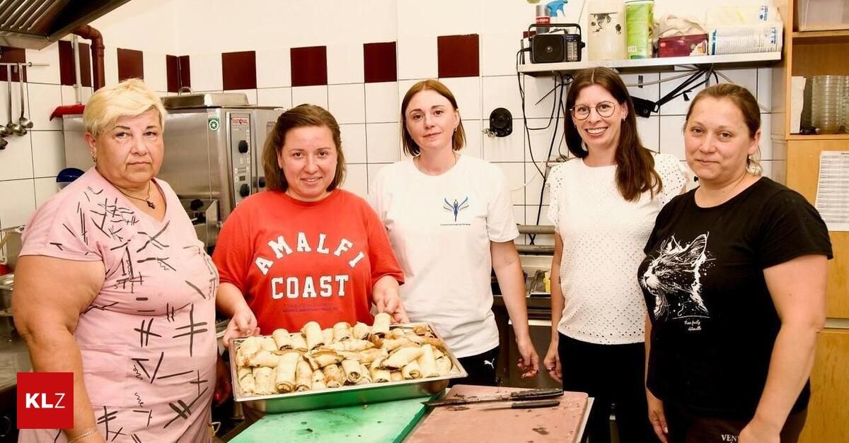 Fünf Frauen in einer Küche, eine hält eine Schüssel mit Essen und lächelt für ein Foto.