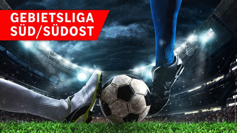 Ein Fußballspieler in blauen Socken und schwarzen Schuhen tritt einen Fußball auf einem Rasenfeld, mit einem verschwommenen Stadion im Hintergrund.