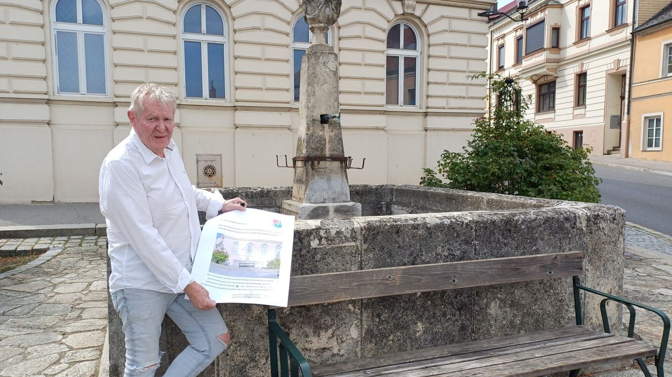 Ein älterer Mann in einem weißen Hemd und Jeans hält ein Papier vor einem Steinbrunnen mit einem Gebäude im Hintergrund.