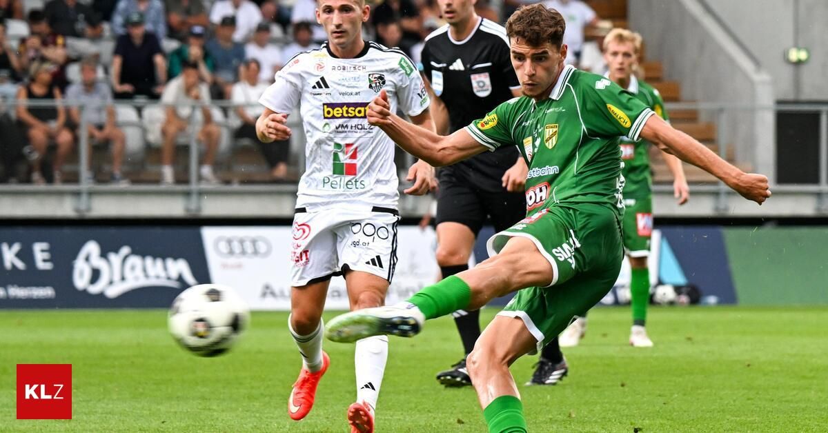 Ein Fußballspieler in einem grünen Trikot tritt den Ball, während ein anderer Spieler in Weiß versucht, den Schuss zu blockieren. Ein Schiedsrichter steht hinter ihnen.
