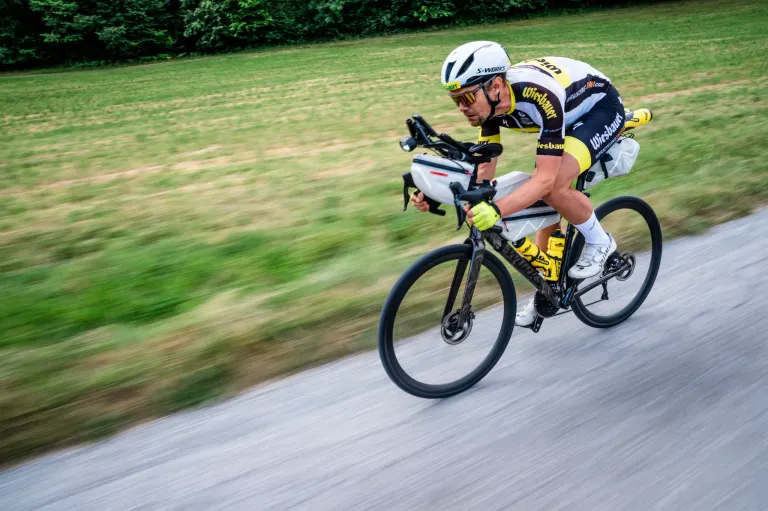 Ein Radfahrer in einem gelben und schwarzen Trikot fährt eine Straße hinunter, mit einem verschwommenen Hintergrund aus grünem Gras.
