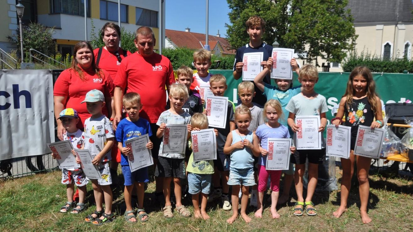 Eine Gruppe von Kindern und Erwachsenen posiert für ein Foto. Die Kinder halten Urkunden in den Händen, und einige Erwachsene tragen rote T-Shirts. Im Hintergrund befindet sich ein Gebäude mit einem roten Dach und einem grünen Zaun.