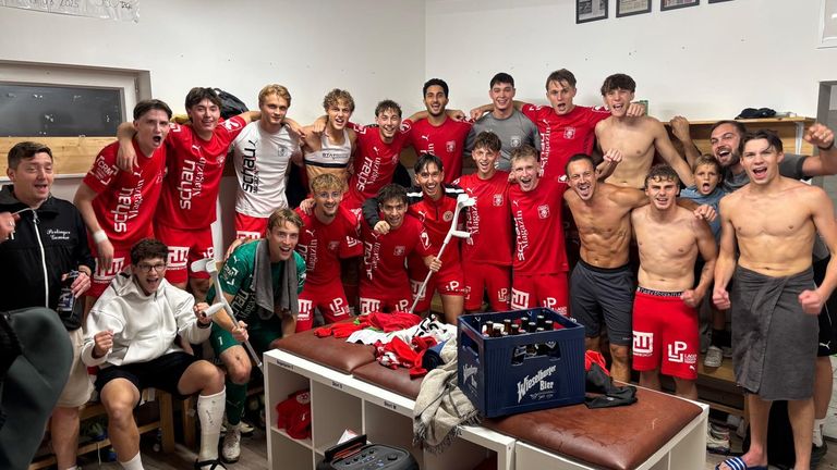 Eine Gruppe junger Männer in roten Uniformen, wahrscheinlich ein Sportteam, posiert für ein Foto in einer Umkleidekabine. Einige von ihnen sind oben ohne, und es gibt eine blaue Kiste Bier auf der Bank.