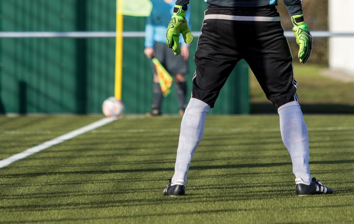 Ein Fußballspieler in schwarz-weißem Trikot mit Handschuhen steht auf dem Spielfeld, während ein Schiedsrichter im Hintergrund eine gelbe Flagge hält.