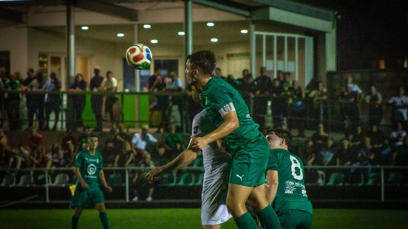 Ein Fußballspieler in Grün springt, um den Ball zu köpfen, während ein anderer Spieler in Grau versucht, ihn zu blockieren. Zuschauer schauen von den Tribünen zu.